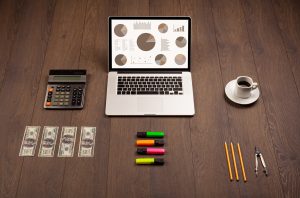 Laptop mit bunten Diagrammen und Statistiken auf Holztisch, umgeben von Taschenrechner, Textmarkern und Kaffee