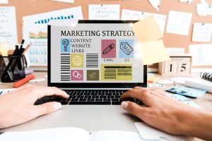 Hände tippen auf einem Laptop, auf dem Bildschirm steht „Marketing Strategy" mit Symbolen für Content, Website, E-Mail und SEO