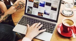 Hände tippen auf einem Laptop mit einer bildlastigen Website auf einem Holztisch, daneben ein Cappuccino und ein Glas Wasser