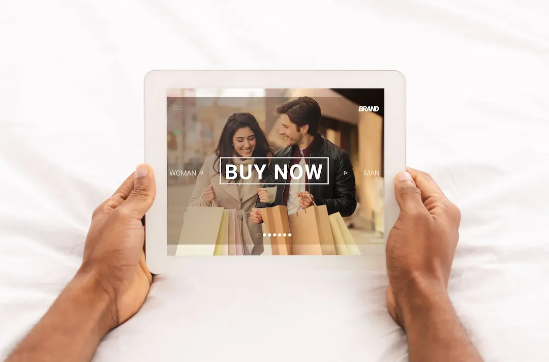 Hände halten Tablet mit Online-Shop und ‚Buy Now‘-Button, Paar mit Einkaufstaschen im Hintergrund.