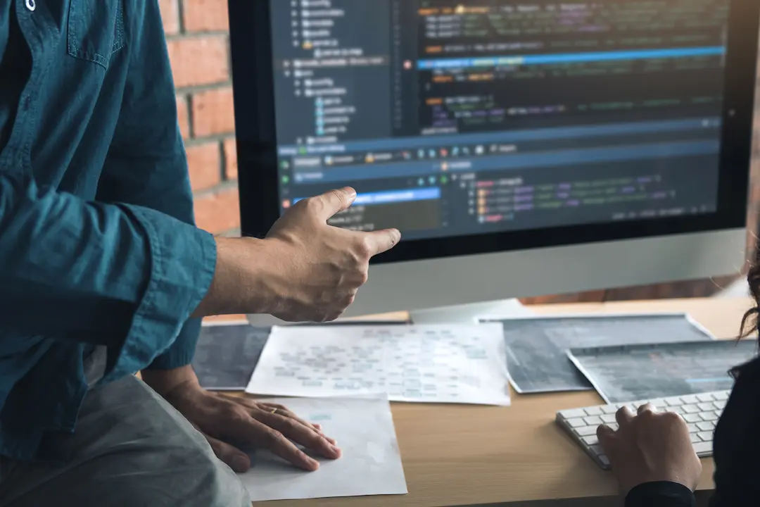Person zeigt auf Code am Monitor, während eine andere am Computer arbeitet.