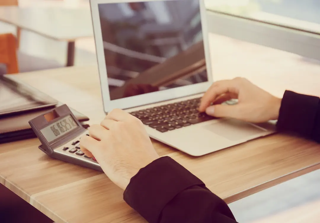 Person nutzt Laptop und Taschenrechner am Schreibtisch.
