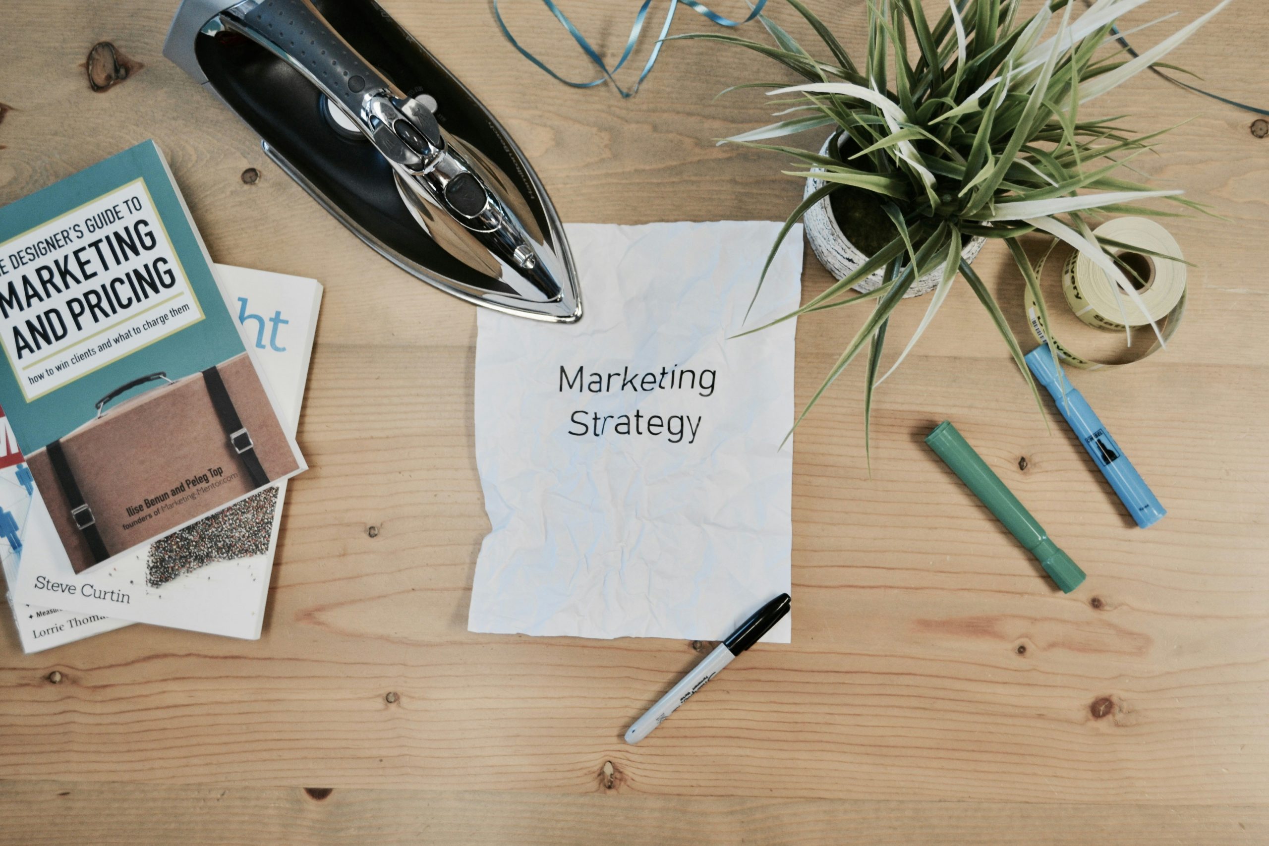 Draufsicht auf einen Holztisch mit einem zerknitterten Blatt Papier mit der Aufschrift „Marketing Strategy", Büchern über Marketing, einem Bügeleisen, Stiften und einer Pflanze