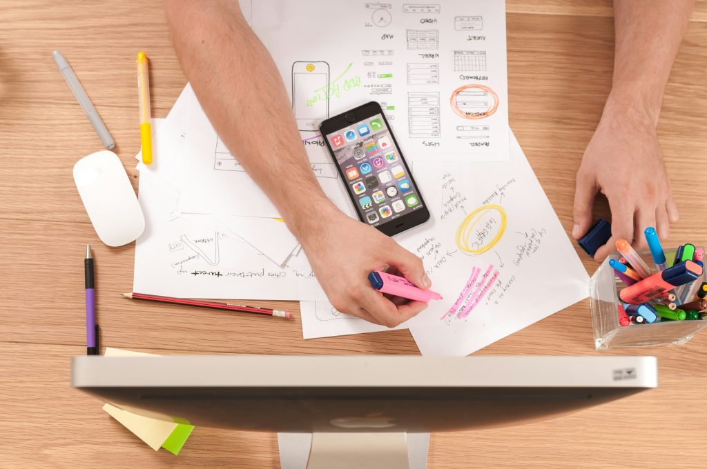 Person arbeitet an UI/UX-Wireframes für eine mobile App – Draufsicht auf einen Schreibtisch mit Smartphone, handgezeichneten Skizzen, Markern und einer Computermaus