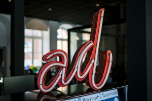 Neon-Schriftzug "ad" in Rot und Weiß auf einer Theke, im Hintergrund ein modernes Büro mit großen Fenstern; davor liegen Exemplare des Bayreuth Magazins