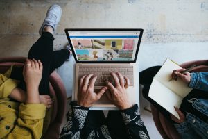 Draufsicht auf zwei Personen bei der Arbeit – eine tippt auf einem Laptop mit Bildstock-Website, die andere macht Notizen in einem Notizbuch