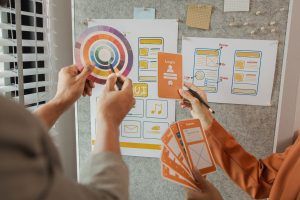 Zwei Designer arbeiten an App-Wireframes mit Farbrad, UI-Skizzen für Login- und Zahlungsbildschirme sowie handgezeichneten Mockups an einer Pinnwand