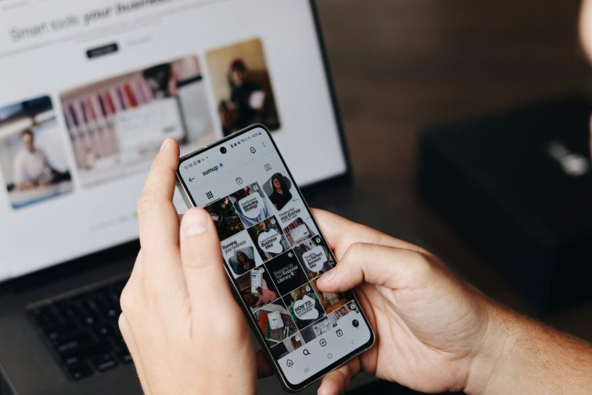 Hände halten Smartphone mit Instagram-Feed vor einem Laptop-Bildschirm