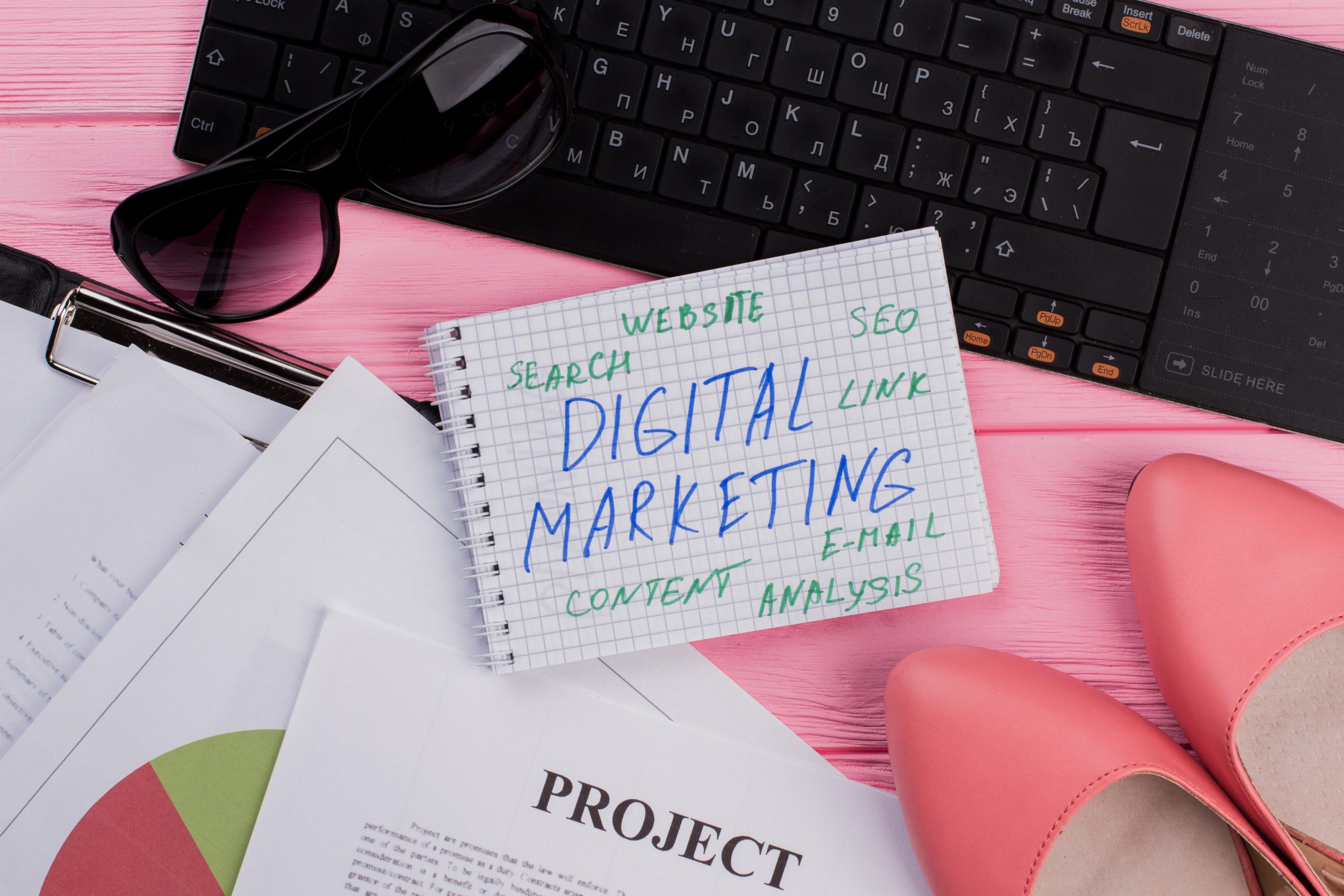 Notizbuch mit handgeschriebenen Begriffen zu Digital Marketing wie SEO, Content und Analyse auf pinkem Schreibtisch