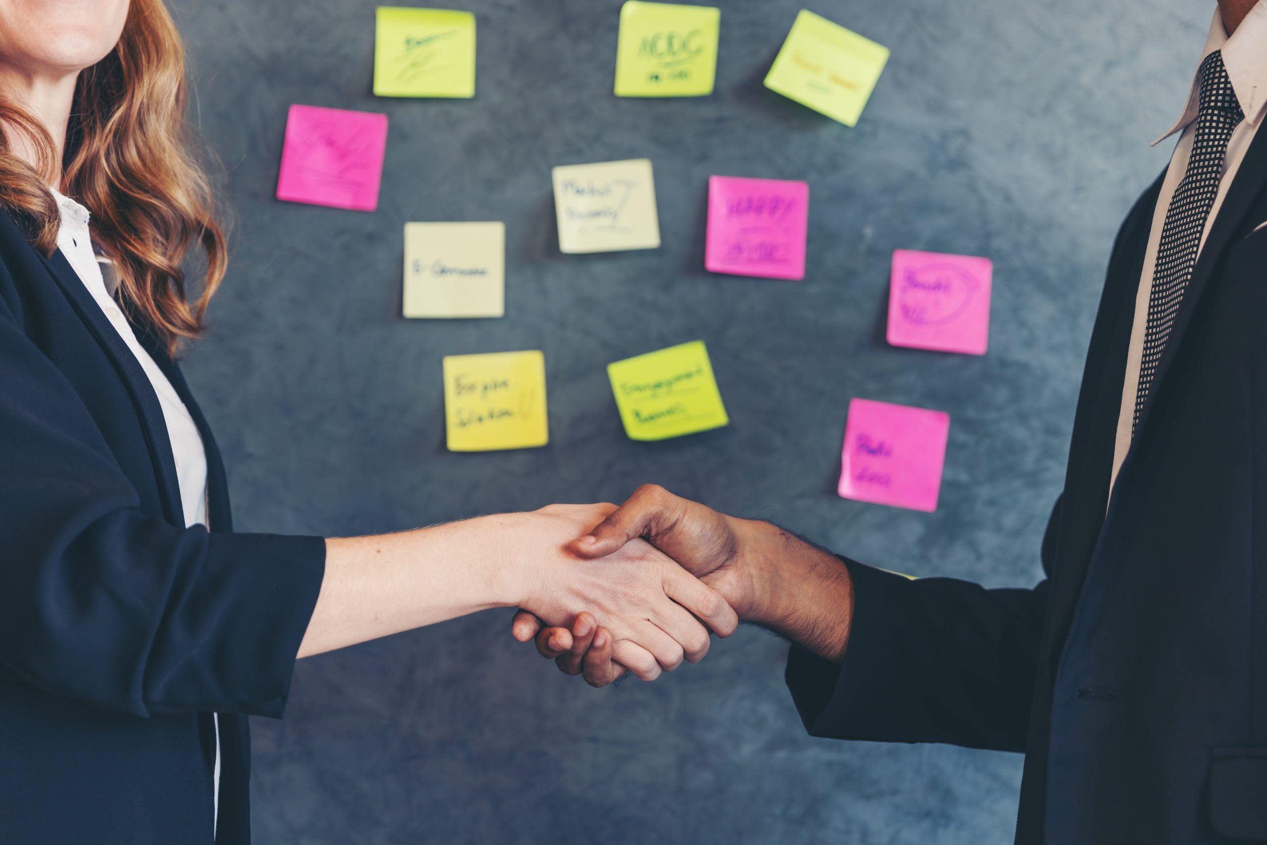 Geschäftspartner geben sich die Hand vor einem Whiteboard mit bunten Notizzetteln zur Projektplanung
