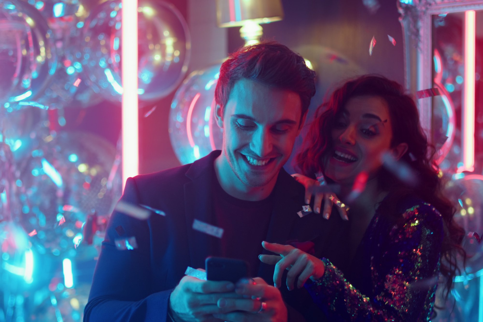 Junges Paar lacht gemeinsam über ein Smartphone bei einer Feier mit Konfetti und Lichterketten