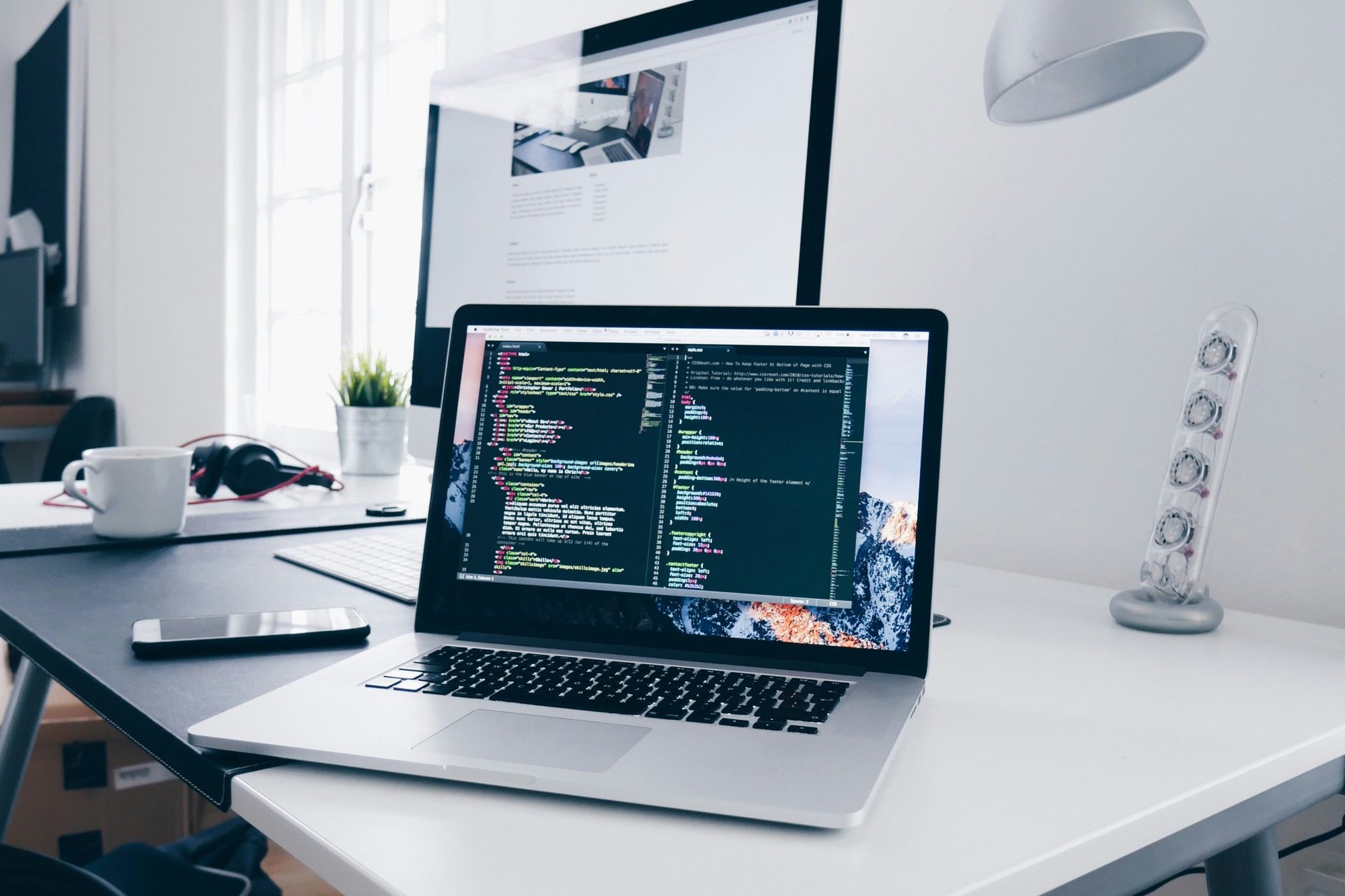Laptop mit Quellcode im Vordergrund und fertiger Website-Vorschau auf externem Monitor