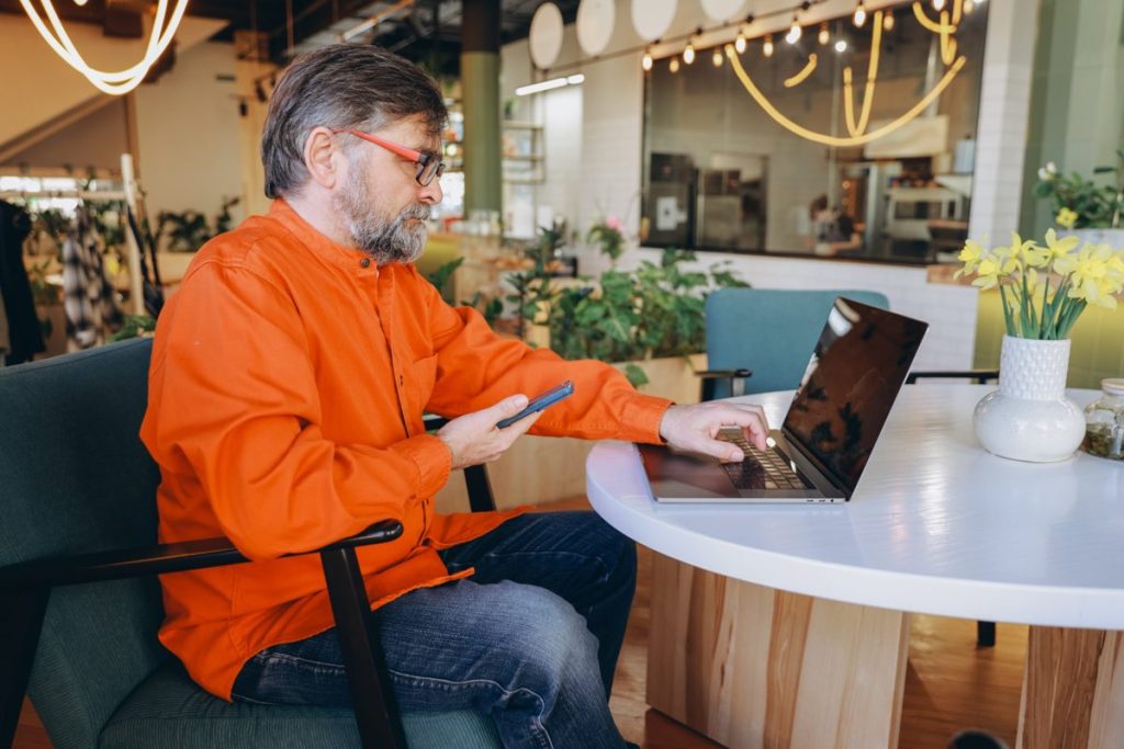 Mann im orangefarbenen Hemd arbeitet im Café am Laptop und hält ein Smartphone, Sinnbild für das Antworten auf Google Bewertungen