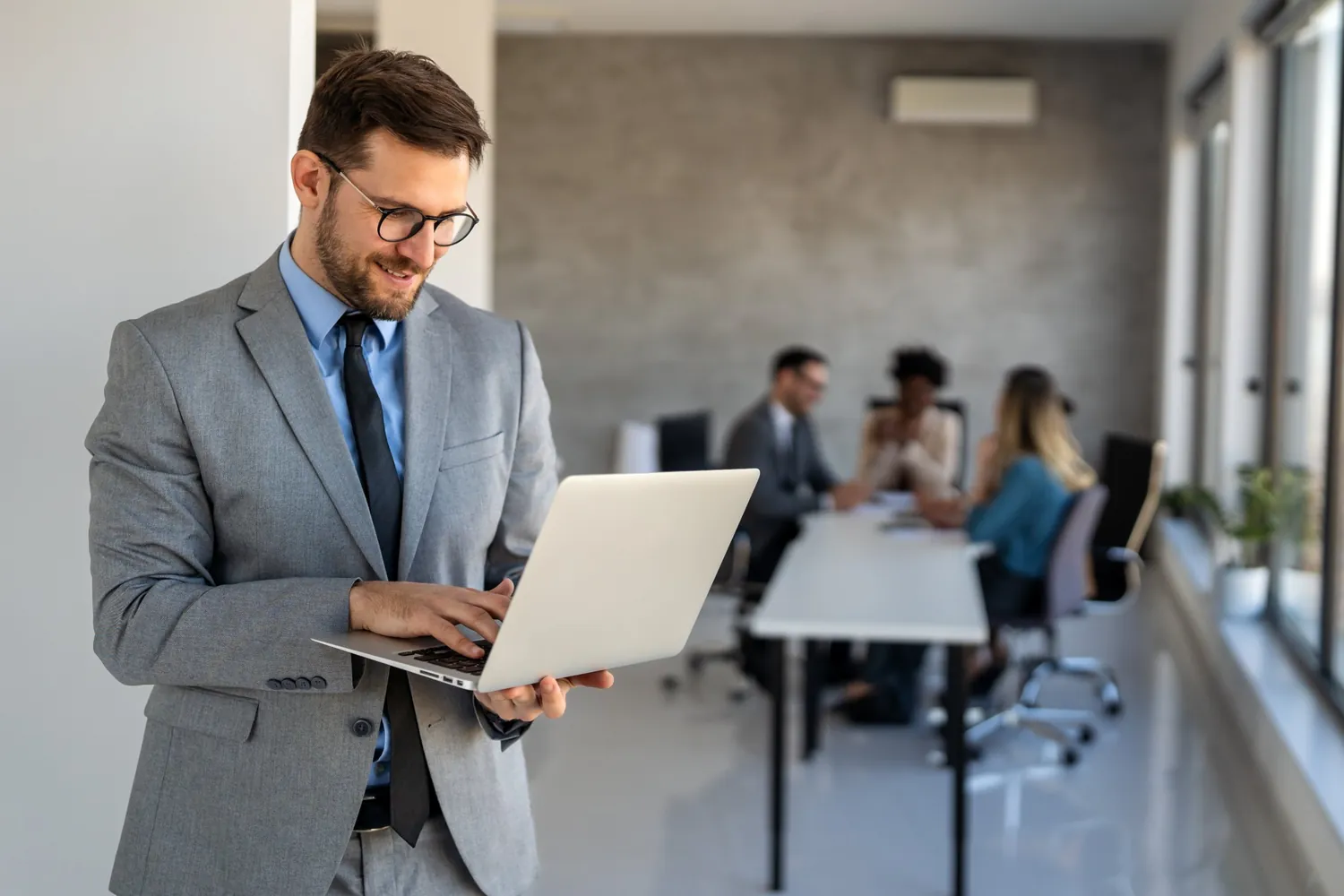 Führungskraft mit Laptop im Büro, im Hintergrund eine Besprechung, Entscheider im B2B erreichen