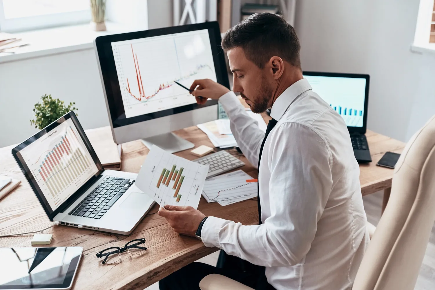 Mann wertet Diagramme auf Monitoren, Laptops und Papier am Schreibtisch aus, Analyse einer Kampagne